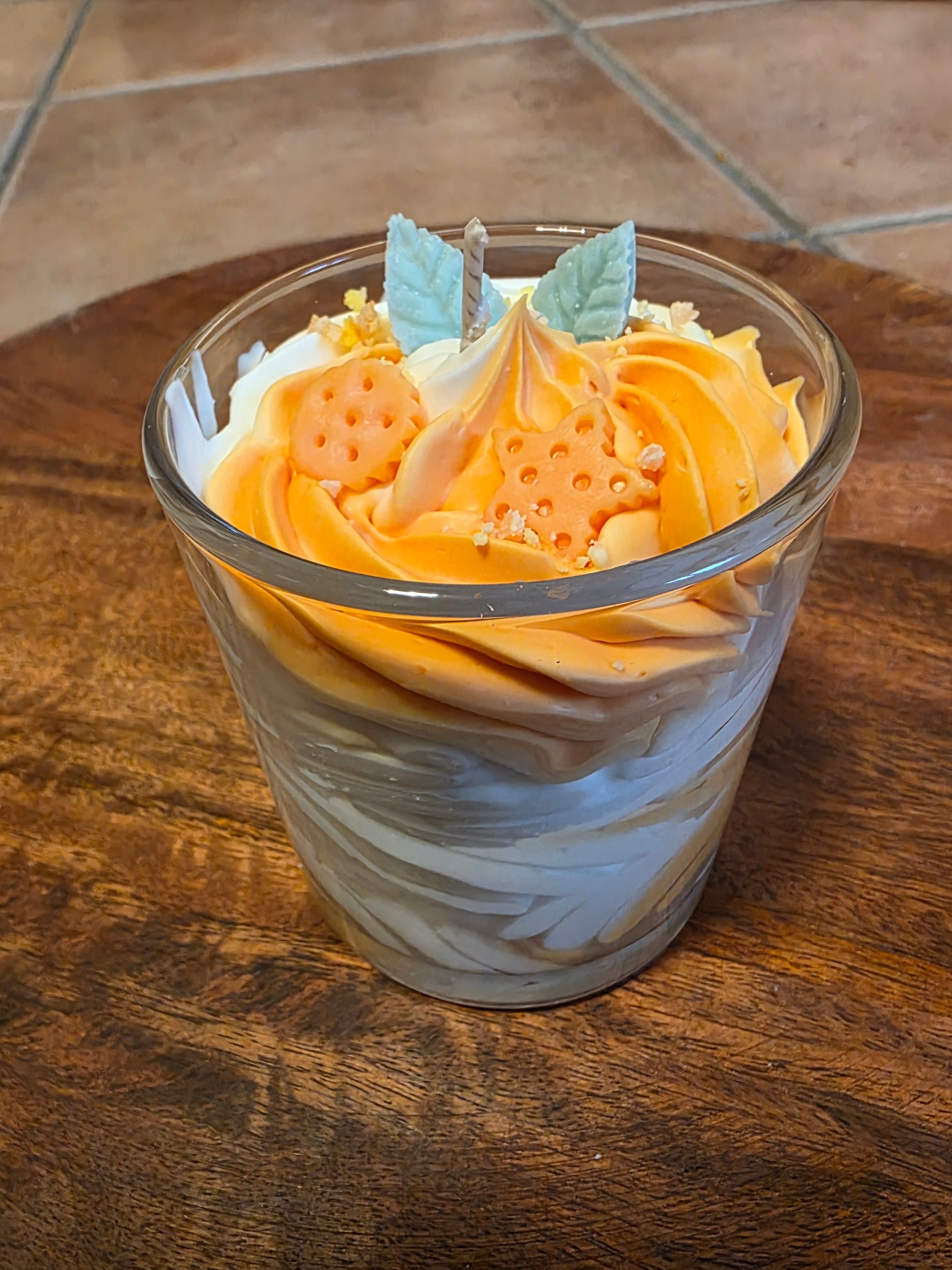 Nahaufnahme der orange-weißen Schichtkerze im Glas, die schräg auf einem runden Holztablett steht, wobei die dekorative Oberfläche mit den Wachsstücken und dem Docht gut sichtbar ist.