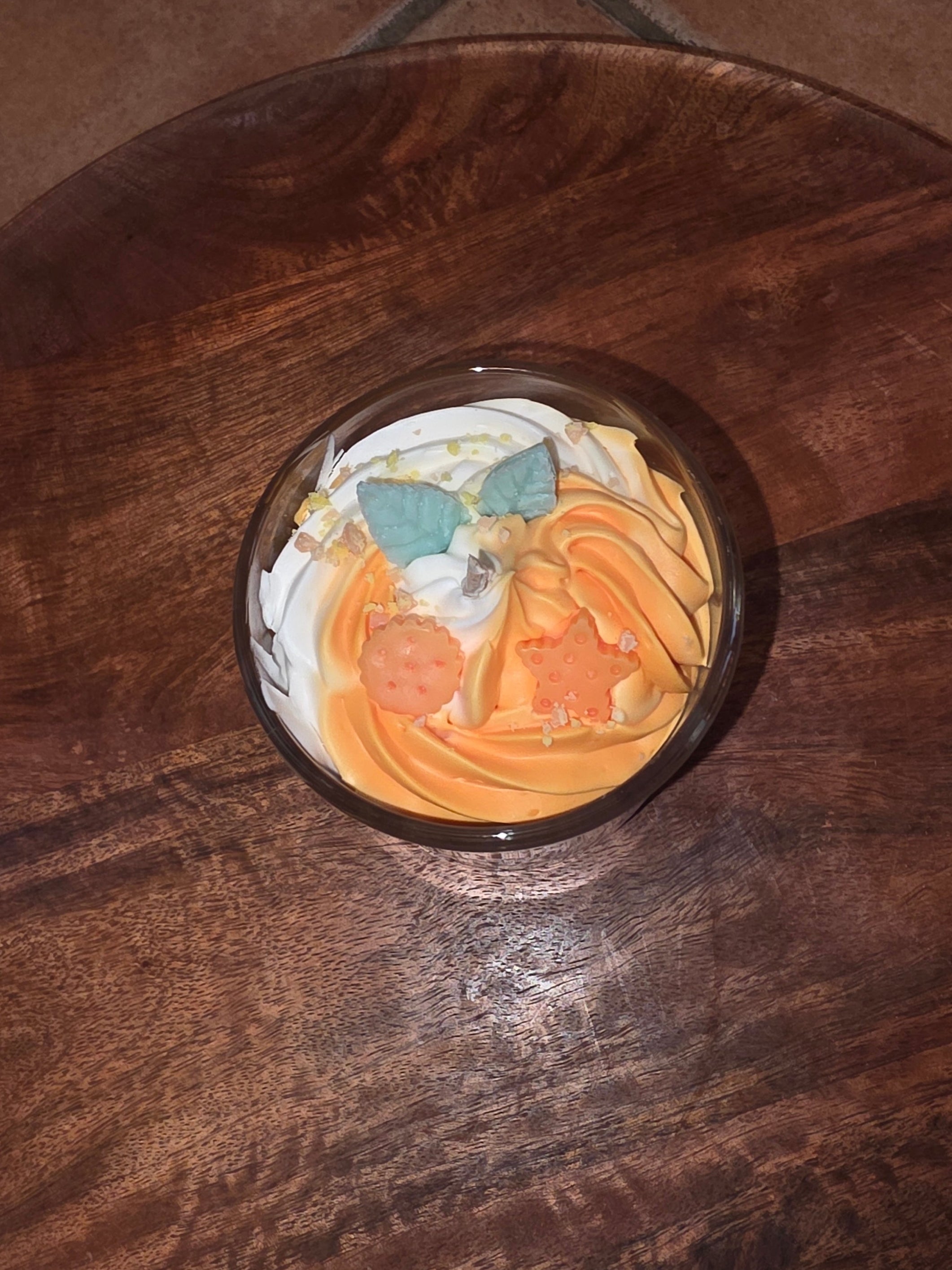 Nahaufnahme der orange-weißen Schichtkerze im Glas, die schräg auf einem runden Holztablett steht, wobei die dekorative Oberfläche mit den Wachsstücken und dem Docht gut sichtbar ist.