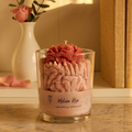 Handgegossene Melina Rose Sojawachskerze von LichtleinLiebe im Glas. Das Dessert-Kerzen-Design zeigt ein rosa Wachs-Blumen-Topping und geschichtete, rosa Wachs-Rüschen. Ein Etikett mit der Aufschrift 'Melina Rose' und 'Sojawachskerze' ist sichtbar. Die Kerze steht auf einem dunklen Holztisch in einem Wohnumfeld.