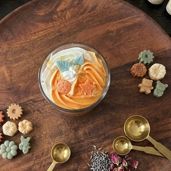 Draufsicht auf ein rundes Holztablett, auf dem eine dekorative orange-weiße Kerze im Glas steht, umgeben von kleinen Seifenstücken in Form von Zahnrädern, Blumen und Teddybären, goldenen Messlöffeln, getrockneten Blüten und verschiedenen Gläsern mit Zutaten im Hintergrund.