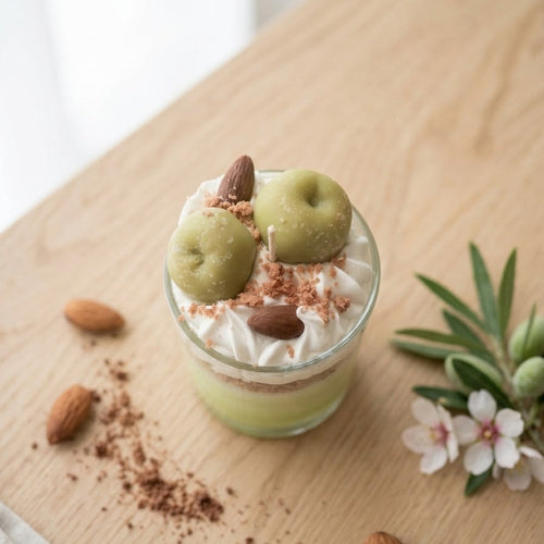 Detailaufnahme von oben der Apfel-Zimt-Crumble Kerze. Zu sehen ist die kunstvoll drapierte Sahnehaube aus Wachs, die mit zwei kleinen, grünen Äpfeln (Wachs-Äpfeln) und zwei braunen Mandeln (Wachs-Mandeln) verziert ist. Eine feine, warme Schicht aus zimtigen Kuchenstreuseln ist über die Sahne gestreut. Im Zentrum der Kerzendocht. Die Aufnahme betont die handwerkliche Qualität und Textur.